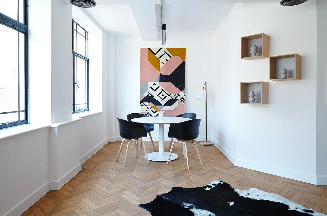 White room with a round dining table, black chairs, wall cubes and a colorful painting