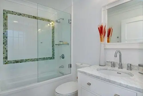 Bathtub with a sliding glass screen and mosaic tile border next to a vanity