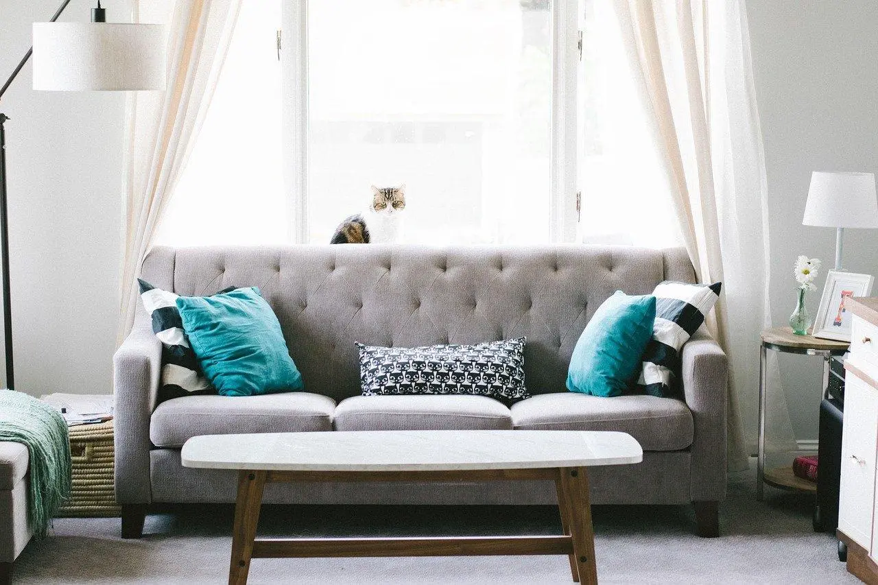 Grey tufted sofa with teal cushions, sheer curtains and a white coffee table